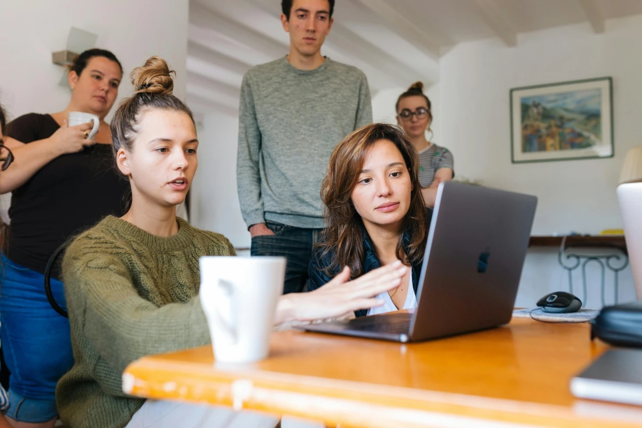 Sechs Personen sitzen und stehen um einen Tisch herum und schauen auf einen Laptop. Eine Person erklärt den anderen gerade etwas.