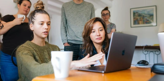 Sechs Personen sitzen und stehen um einen Tisch herum und schauen auf einen Laptop. Eine Person erklärt den anderen gerade etwas.